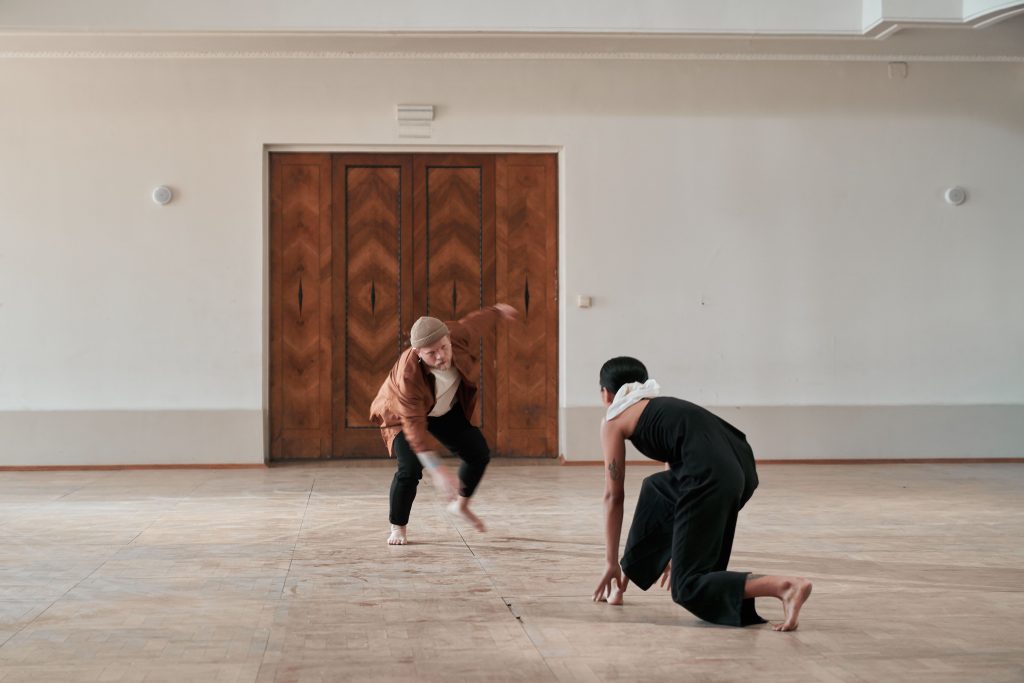 Two dancers performing contemporary dance in a minimalist indoor setting.
